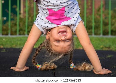 Little Girl Stands On Her Hands And Smiles. Child Has Fun Outside. Kid Enjoys Upside Down. Exercises, Coups, Tumbling, Standing Upside Down, Laughter And Happiness. Happy Childhood Concept. 