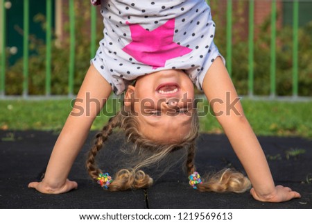 Little girl stands on her hands and smiles. Child has fun outside. Kid enjoys upside down. Exercises, coups, tumbling, standing upside down, laughter and happiness. Happy childhood concept. 