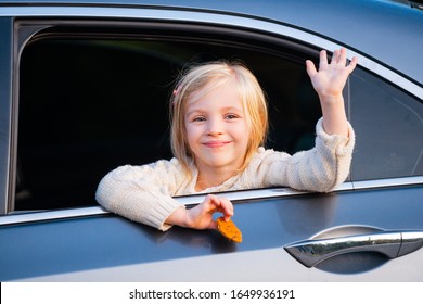 A Little Girl Is Sticking Her Head Out The Car Window. Road Trip Or Travel Concept. Happy Kids Travel By The Car. Cute Child Leaning Out A Van Window, Smiling And Waving