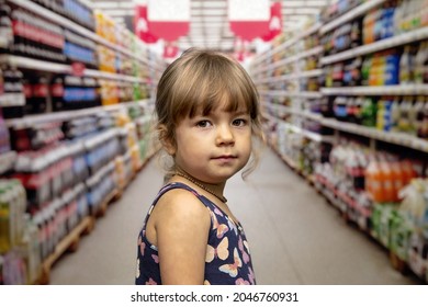 A Little Girl In A Summer Dress On A Blurry Background In A Supermarket. Concept: A Child Got Lost In A Store, Shopping For Children, A Large Selection Of Children's Products.