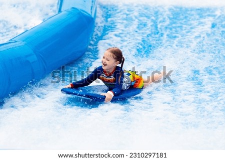 Little girl surfing in beach wave simulator attraction in water amusement park of tropical resort. Child trying to ride body board. Kids surf. Preschooler kid surfer during training on generated waves