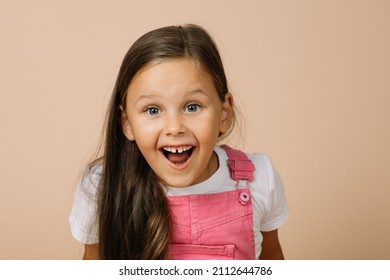 Little girl with surprised eyes, opened mouth, blissful smile with teeth and raised eyebrows looking at camera wearing bright pink jumpsuit and white t-shirt on beige background. - Shutterstock ID 2112644786