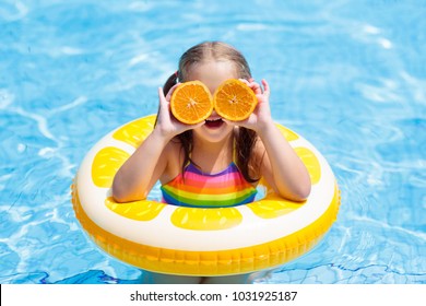 Little Girl In Swimming Pool With Inflatable Toy Ring Eating Orange. Kids Swim On Summer Vacation. Tropical Fruit And Healthy Snack. Swim Aids For Child.  Kid On Colorful Float. Beach And Water Fun.