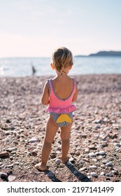 Little Girl In A Swimsuit Stands On The Beach. Back View