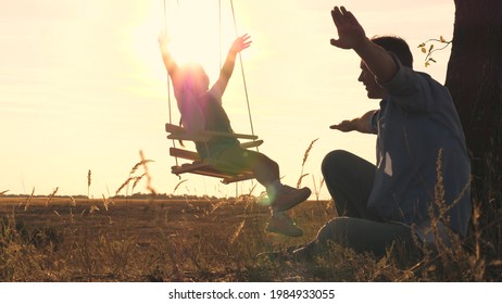 Little Girl Swinging On A Swing At Sunset And Waving Her Arms In Flight, Soar Like An Airplane Above The Ground, Slow Motion, Happy Family, Teamwork, Playing With A Child Outdoors Together