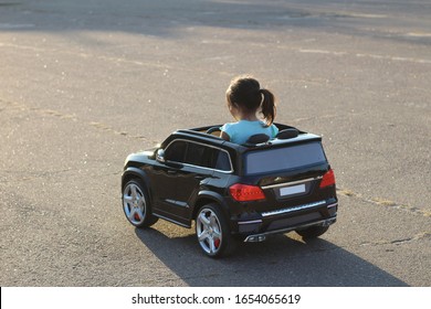little girl with a tail drives a toy black car on asphalt in a square in the city