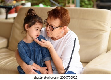 Little Girl Talking To Her Grandma Sitting On The Couch. Portrait Of Loving Grandmother And Granddaughter Together At Backyard. Little Child Is Sitting On Grand Mom’s Lap.