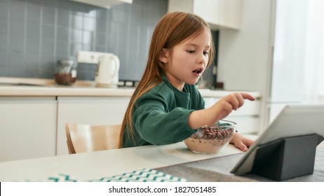 A Little Girl Is Tapping On A Tablet Screen While Eating Her Quick Cereal Balls With Milk From A Glassy Bowl Sitting At The Table In A Big Bright Kitchen, Child Spending Time At Home