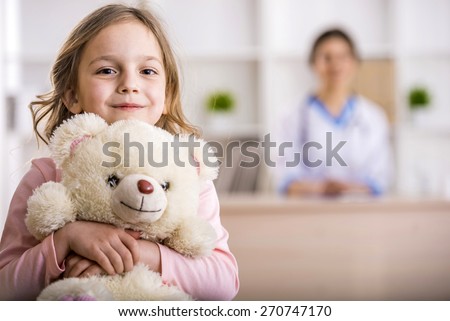 Little girl with teddy bear is looking at the camera. Female doctor on background.