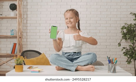 Little girl toddler sitting on the table in living room, child holding smartphone with chroma key green screen background workspace mockup shows thumb up. - Powered by Shutterstock