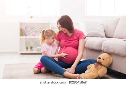 Little girl touching her pregnant mom belly. Expecting mother sitting with her daughter on floor at home, having fun and feeling baby pushes, copy space