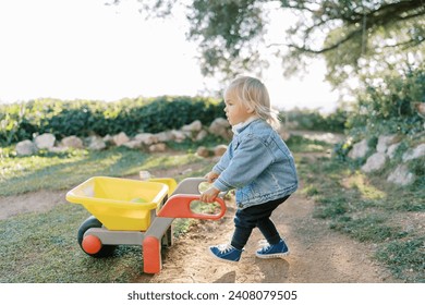 Little girl with a toy wheelbarrow walks along a dirt path in the park - Powered by Shutterstock