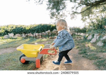 Little girl with a toy wheelbarrow walks along a dirt path in the park