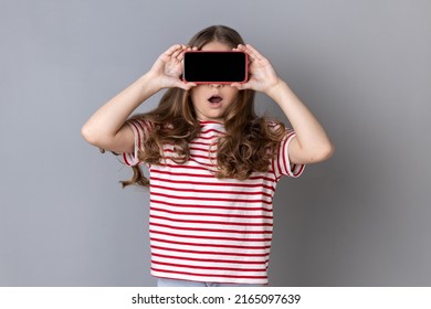 Little Girl In T-shirt Covering Eyes With Cellphone, Posing With Mouth Wide Open From Shock Surprise, Unknown Child Hiding Face With Mobile Phone. Indoor Studio Shot Isolated On Gray Background.