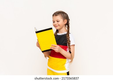 A little girl in a T-shirt with a German flag reads a yellow book on a white isolated background.