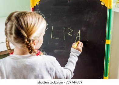 little girl with two pigtails is  writing her name with chalk on a blackboard, with copy space - Powered by Shutterstock