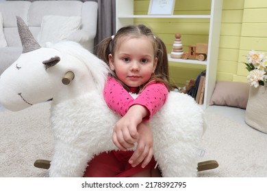 Little girl with a unicorn in the children's room. Child 4-5 years old in a children's room with toys. A baby in a pink dress is sitting on the floor near a white pony. The child plays alone in the