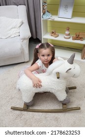 Little girl with a unicorn in the children's room. Child 4-5 years old in a children's room with toys. A baby in a white dress is sitting on the floor near a white pony. The child plays alone in the