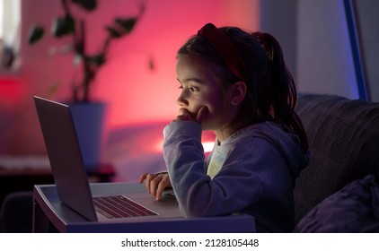 A Little Girl Uses A Laptop Late At Night, A Child Sits In Front Of A Computer Screen In The Room With Neon Lightning, The Concept Of Gadget Addiction.