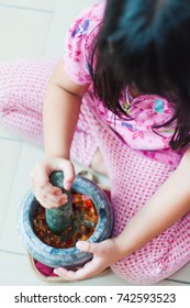 A Little Girl Is Using The Mortar And Pestle Or Lesung Batu In Malay With Crushed Chilies, Fried Shallots And Shrimp Paste Mixed Together. Soft Focus, Shallow Depth Of Field.