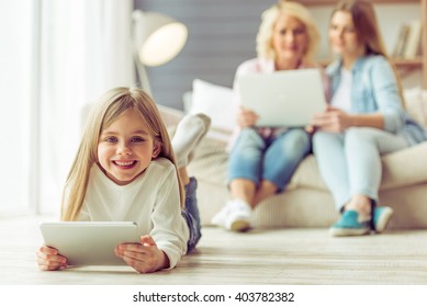 Little Girl Is Using A Tablet, In The Background Her Mom And Granny Are Using A Laptop While Sitting On Sofa At Home