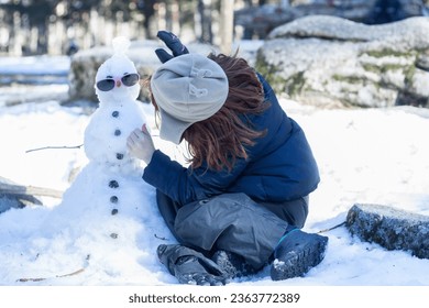 little girl in warm clothes playing with snowman in snowy forest - Shutterstock ID 2363772389