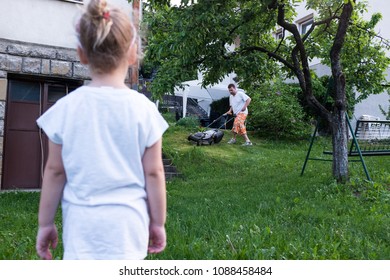 Little Girl Watching Daddy Mow The Lawn In The Backyard, Sunday Afternoon Family Activity, Carefree Childhood Growing Up Coming Of Age Concept