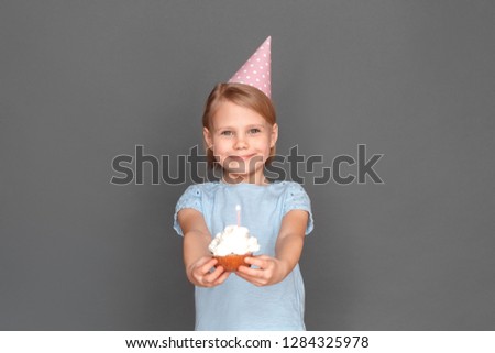 Little girl wearing birthday cap standing isolated on grey wall giving cupcake with lighted candle to camera smiling cheerful
