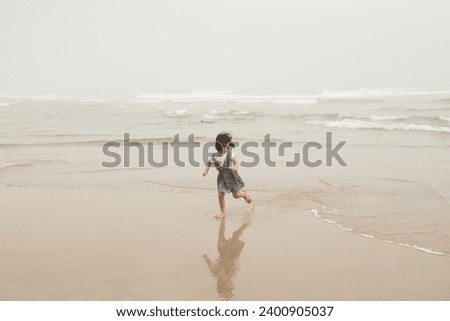 A little girl wearing a dress runs away from the waves as she looks back at the water in a misty day in Playa de las Catedrales (Cathedrals Beach) in Ribadeo, Galicia, Spain
