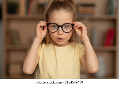 Little Girl Wearing Eyeglasses Having Poor Eyesight Posing Looking At Camera Through Eyewear Standing Near Bookshelf At Home. Kid's Eyes Health. Knowledge And Reading Concept - Shutterstock ID 2154549439