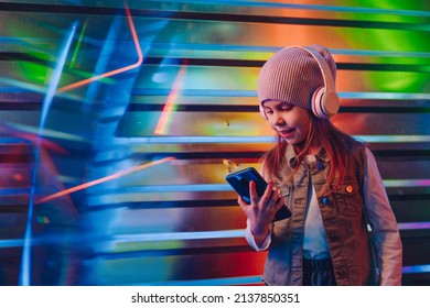 Little girl wearing headphones using smartphone standing on the colorful neon lights background with blank space for text. Generation Alpha using technology. Selective focus on the face.