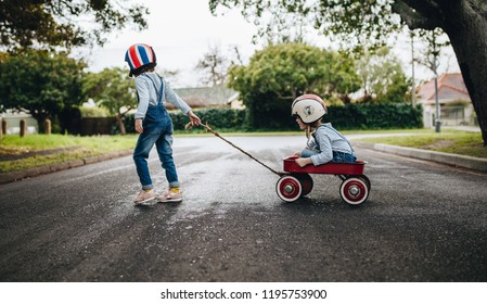 Little girl wearing helmet pulling her sister sitting in a wagon cart on the road. Kids playing outdoors with toy trolley.