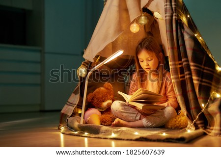 A little girl wearing pyjamas sitting on a floor cross-legged barefoot in a self-made hut made of a plaid, reading a book aloud with her teddybear and fancy garlands all around her spacy room at home