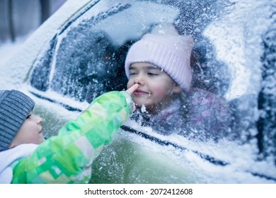 Little Girl Wearing In Winter Clothes Pressed Her Face Against Car Window Glass Funny Flattening Nose And Boy Points Her Nose With Finger, Family Road Trip In Winter, Active Weekend