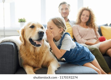 Little girl whispering something to her pet while relaxing on sofa on background of young couple