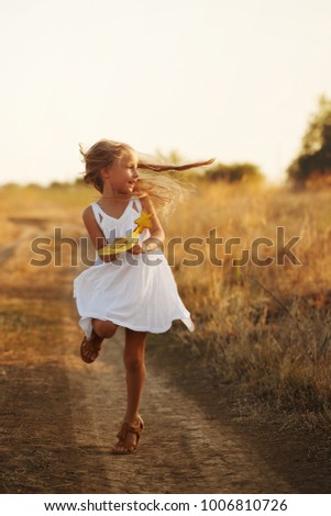 A little girl in a white dress and sandals runs along a country road. She holds a toy in her hands. Happy childhood. Summer vacation