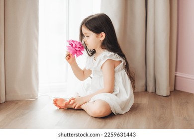 A little girl in a white dress is sitting near the window and holding a rose. A happy child at the window with a flower.