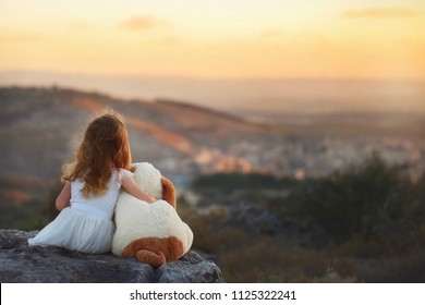 Little girl in white dress with toy friend looking forward on beautiful sunset