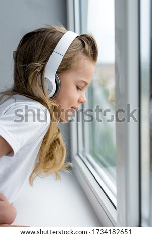little girl with white hair and in a white T-shirt against a gray wall listens to music in white big headphones looks out the window