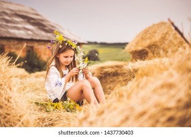 LIttle Girl In White Shirt Sitting On A Haystack And Making A Flower Wreath