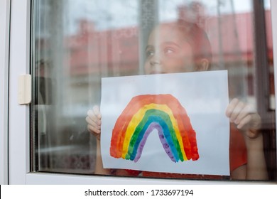 Little girl in the window holds a picture of a rainbow symbolizing the imminent end of the quarantine in connection with the coronavirus pandemic. - Powered by Shutterstock