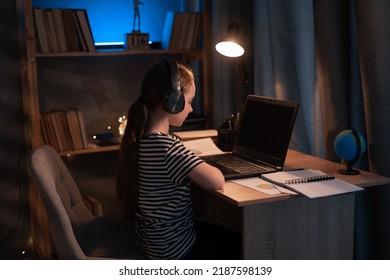 Little Girl In Wireless Headset Using Laptop, Studying Online At Home At Night, Interested Happy Student Typing On Keyboard Looking At Pc Screen, Watching Webinar, Online Course, Doing Homework