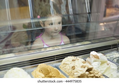 Little Girl Wishes Ice Cream In Pastry Shop. Reflections On Ice Cream Counter Glasses