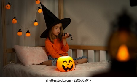 Little Girl In Witch Black Hat Plays With Spider In Dark With Orange Pumpkin Lantern. Happy Child Celebrats Halloween