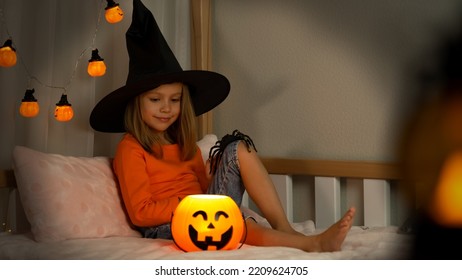 Little Girl In Witch Black Hat Plays With Spider In Dark With Orange Pumpkin Lantern At Home. Happy Child Celebrats Halloween