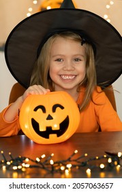 Little Girl In Witch Costume And Black Hat Looks At Camera And Smiles. Happy Child Celebrats Halloween With Lantern Pumpkin