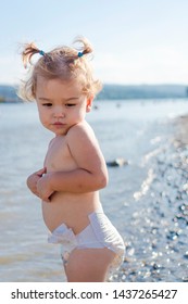 Little Girl With Worried Face Standing Outdoors On Beach Shore. Kid Afraid Of Water. Child With Pigtails Cold On Sunny Day. Sad Toddler In Diapers Looking At River On Windy Day. 