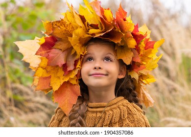 A little girl with a wreath of maple leaves on her head in a warm sweater on an autumn day.