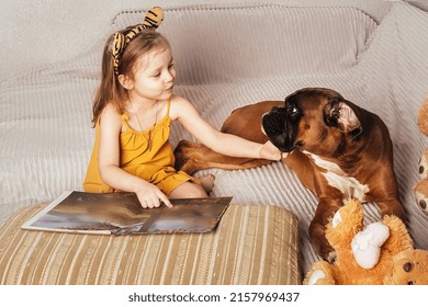 A Little Girl In A Yellow Dress And Wavy Hair Reads A Book And Shows Pictures To Her Big Red Dog On The Sofa At Home