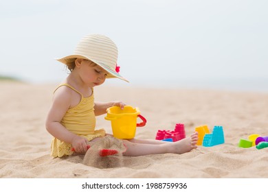Little girl in yellow swimsuit and straw hat sitting on sand and playing with bucket and spade at sea beach in warm sunny summer day. Closeup. Side view.